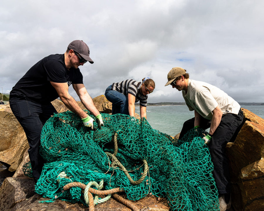 Three people working with a large green fishing net on rocks by the sea.