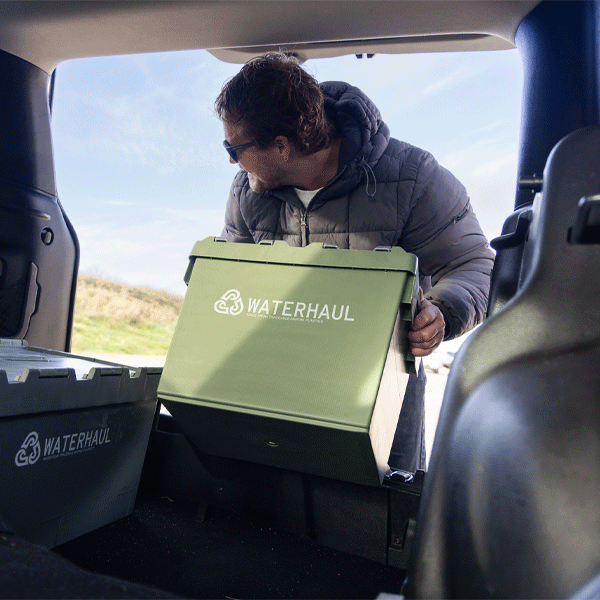 Person loading a green WATERHAUL storage box into a vehicle with a scenic background.