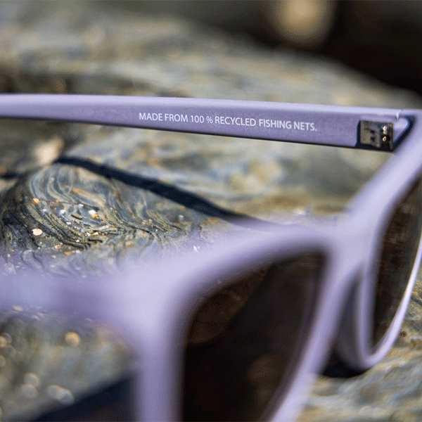 Purple sunglasses with a label on the frame indicating recycled fishing nets material, placed on a textured surface.