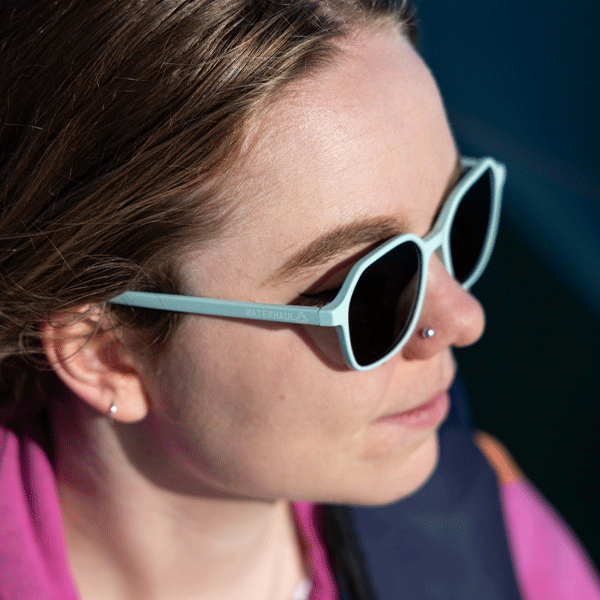 Woman wearing light blue sunglasses and pink jumper with water in background. 