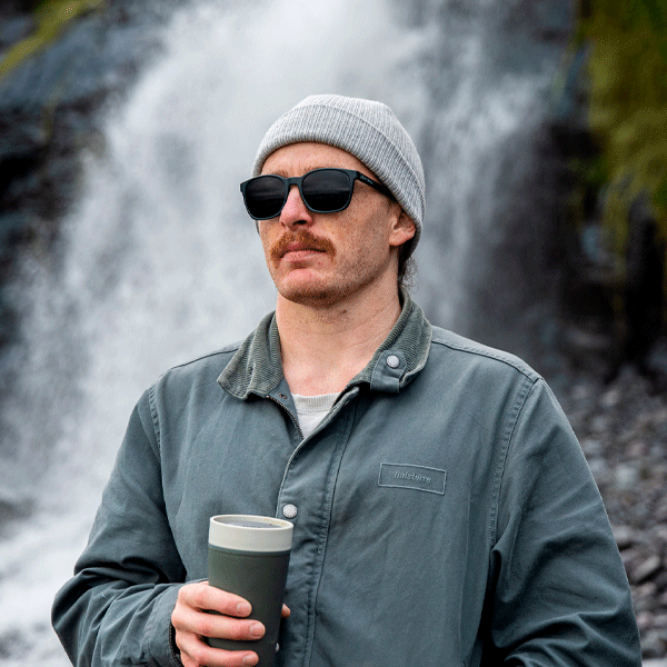 Man wearing black sunglasses and a beanie, holding a coffee cup, with a waterfall in the background