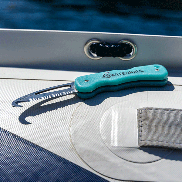 Green pocket knife with a hooked serrated blade on the side of. boat with water in the background