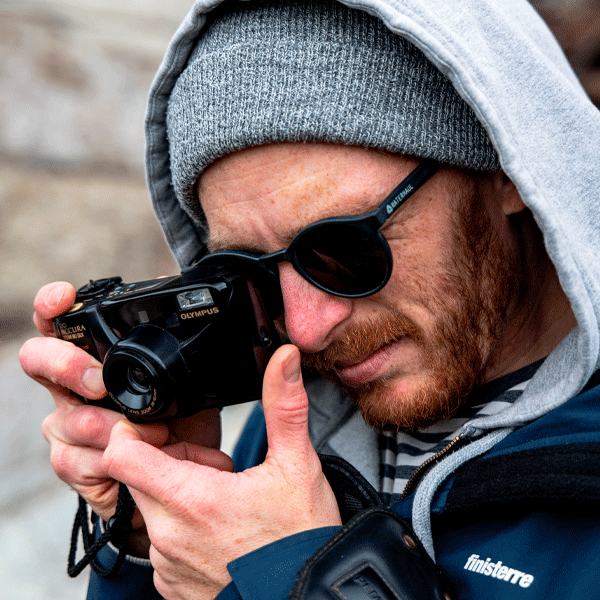 Man wearing round sunglasses and a hooded jacket holding an Olympus camera.
