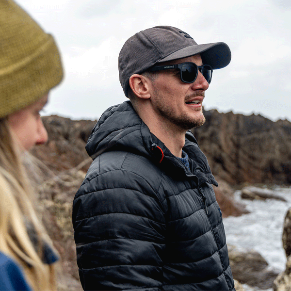 Man wearing a black puffer jacket and sunglasses standing outdoors with a rocky coastal background