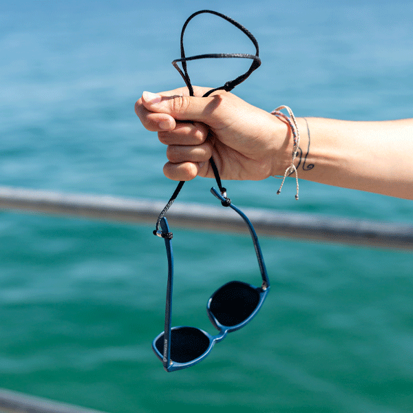 Hand holding sunglasses with a black strap against a water background