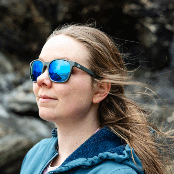 Person wearing sunglasses with a reflective blue lens, standing against a natural rocky background.